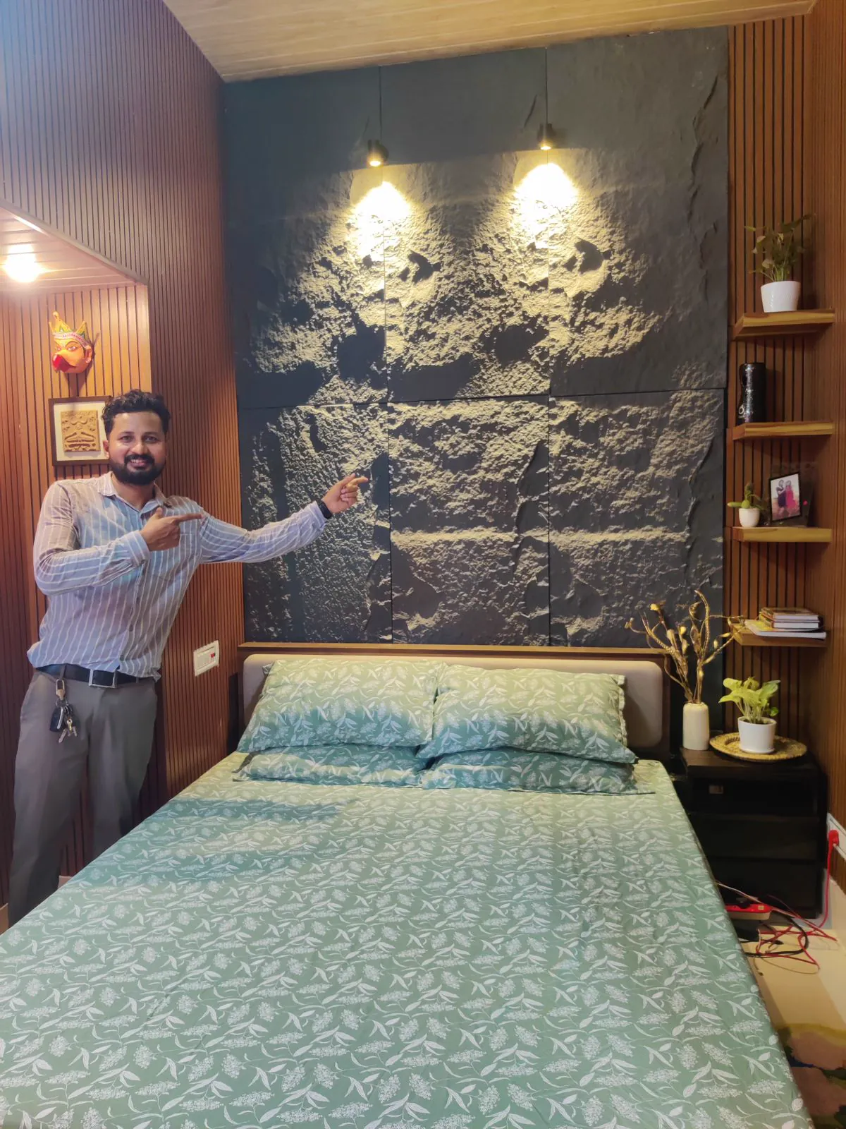 Bedroom with a dark riven stone-effect panel behind the bed, two picture lights, slat walls and open shelving